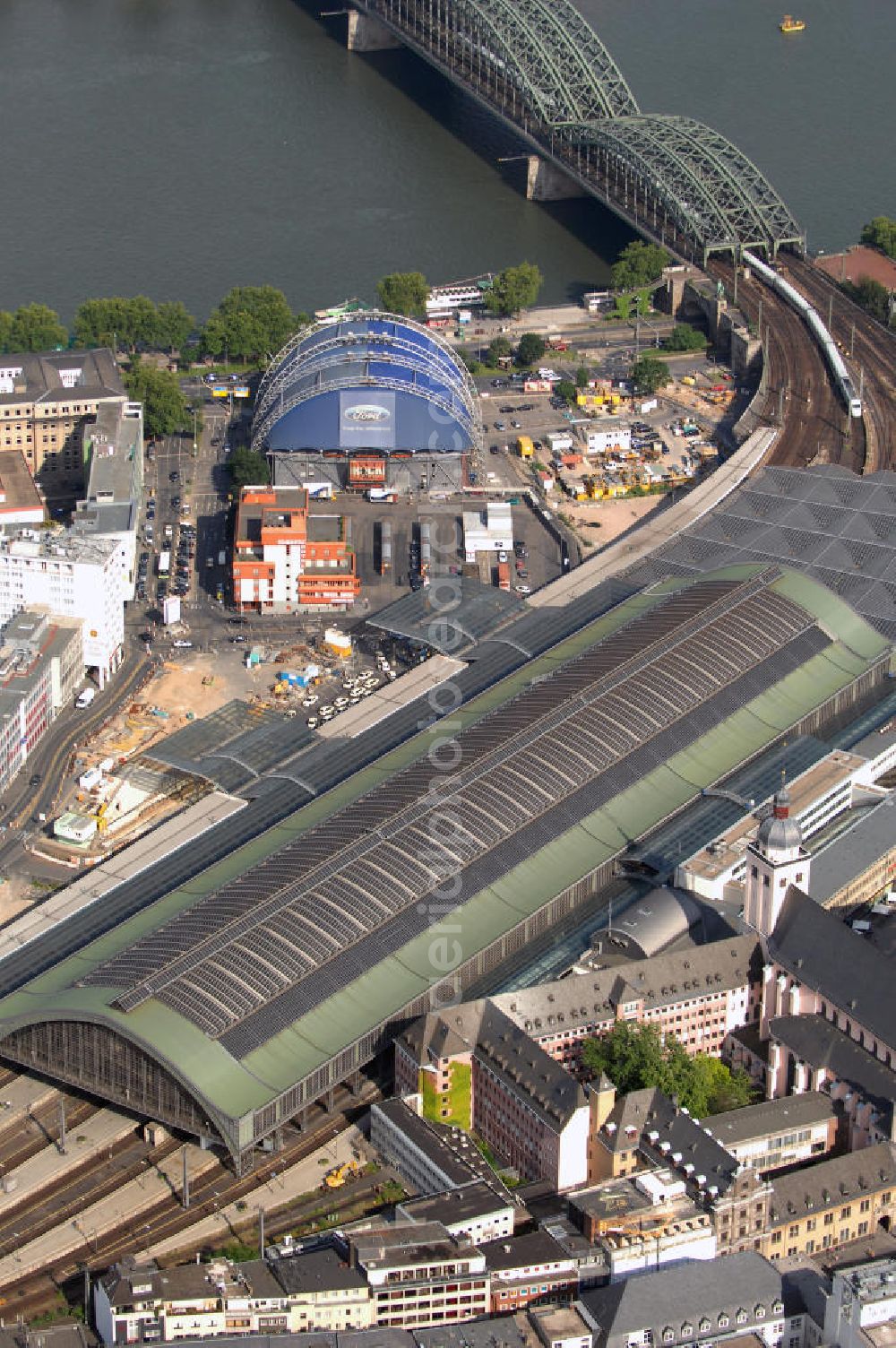 Aerial photograph Köln - Blick auf den Kölner Hauptbahnhof am Fuße des Kölner Doms im Stadtzentrum. Er gehört zu den verkehrsreichsten Bahnhöfen Deutschlands und ist wichtiger Knotenpunkt des deutschen Personenverkehrs. Der Kölner Hauptbahnhof ist ein Durchgangsbahnhof mit elf Gleisen, was für einen solchen Knotenbahnhof wie er es ist, verhältnismäßig klein ist. Um den täglich 250.000 Reisenden die Wartezeiten zu verkürzen, gibt es 71 Geschäfte und Restaurants in der Bahnhofshalle, sowie Schließfächer für das Gepäck oder auch Fahrrad-Stellplätze. Täglich fahren am Kölner Hauptbahnhof 243 Züge des Fernverkehrs und 521 Züge des Nahverkehrs, hinzu kommen 466 S-Bahnen am Tag. Hinter dem Hauptbahnhof ist das Theater Musical Dome an der Rheinuferpromenade zu sehen. Die Eröffnung des Hauses fand 1996 nach nur sechsmonatiger Bauzeit statt. Die außergewöhnliche Glas-Stahl-Bauweise macht es zu einem einzigartigen Theater, das die Blicke auf sich zieht. Der Musical Dome präsentiert sowohl Musicals mit einer Spieldauer von mehreren Jahren wie z.B. Saturday Night Fever oder auch das noch aktuelle Queen Musical, als auch kürzere Shows wie den chinesischen Nationalzirkus, Buddy Holly oder die Show des Magiers David Copperfield. In der Mitte des oberen Bildrandes sieht man die Hohenzollernbrücke. Sie führt über den Rhein und bestand ursprünglich aus zwei Eisenbahn- und einer Straßenbrücke. Die Hohenzollernbrücke löste 1911 die Dombrücke ab, die zuvor an der Stelle war, dem wachsenden Verkehr jedoch nicht mehr standhielt. Heute existiert die Straßenbrücke der Hohenzollernbrücke nicht mehr, sie wurde ebenfalls durch eine Eisenbahnbrücke ergänzt und die gesamte Brücke links und rechts durch Fußgänger- und Radwege erweitert. Kontakt Veranstalter Musical Dome: TKS Ticket-Service und Veranstaltungen GmbH, Erkrather Straße 30 40233 Düsseldorf, Tel. +49(0)211 7344 0, Fax +49(0)211 7344 155, Email: webmaster@musicaldome.de