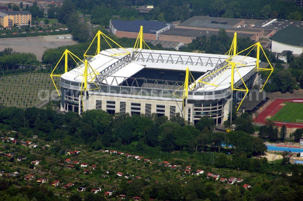 DORTMUND from above - Der Signal Iduna Park (bis Ende November 2005 Westfalenstadion) ist mit 80.708 Zuschauerplätzen in Bundesliga-Konfiguration das größte reine Fußballstadion Deutschlands. Er ist die Spielstätte des Fußball-Bundesligisten Borussia Dortmund und war bereits mehrfach Austragungsort internationaler Fußballturniere, zuletzt der Fußball-Weltmeisterschaft 2006, sowie von Heimspielen der deutschen Nationalmannschaft. Das Stadion mit den markanten gelben Pylonen der Dachkonstruktion ist ein Wahrzeichen der Stadt Dortmund. In der zweiten Ausbaustufe wurden auch Nord- und Südtribüne mit einem Oberrang überbaut. Auf der Nordtribüne wurden im Oberrang Sitzplätze errichtet, während der Oberrang der Südtribüne traditionell als Stehplatz realisiert wurde. Als reine Stehplatztribüne fasst die Südtribüne seitdem fast 25.000 Zuschauer (exakt: 24.454) und ist damit die größte in Europa; insgesamt bot das Stadion nach diesem Ausbau 68.600 Besuchern Platz. Bei internationalen Spielen kann die Stehplatztribüne innerhalb kurzer Zeit mit Sitzplätzen bestückt werden. Die Zuschauerkapazität des Stadions verringert sich dabei um rund 15.000 Plätze. Die erste Ausbaustufe wurde von der Hochtief AG durchgeführt, die zweite vom Osnabrücker Baukonzern Köster AG. Zwischen den Jahren 2002 und 2003 wurde die dritte Ausbaustufe durchgeführt. Ziel war es, die bislang offenen Stadionecken zu schließen und damit die Kapazität des Stadions weiter zu erhöhen. Planende Architekten waren das Architekturbüro Schröder Schulte-Ladbeck, die Tragwerksplanung übernahm die Firma Engels Ingenieure GmbH, Berger & Berger.Nach der dritten Ausbaustufe belief sich die Kapazität des Stadions auf knapp 83.000 Zuschauer. Die Außenseite der Nordtribüne wurde verglast, im Inneren finden sich Gastronomiebereiche. Auffälliges bauliches Merkmal sind die außengeführten, leuchtend gelben Stahlpylonen, die weit über das Dach hinausragen und von vielen Punkten der Stadt sichtbar sind.