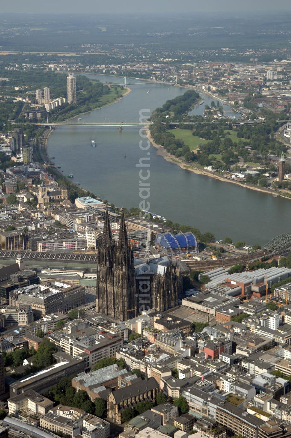 KÖLN from the bird's eye view: Der Kölner Dom, offizieller Name Hohe Domkirche St. Peter und Maria, ist eine römisch-katholische Kirche in Köln und die Kathedrale des Erzbistums Köln.Der Kölner Dom ist mit 157 Metern Höhe nach dem Ulmer Münster die zweithöchste Kirche in Deutschland sowie die dritthöchste der Welt. Er steht an der nördlichen ehemaligen römischen Stadtgrenze in direkter Nachbarschaft des heutigen Hauptbahnhofs, der Altstadt, Hohenzollernbrücke und Museum Ludwig und ist von einer modernen Betonkonstruktion, der sogenannten Domplatte, umgeben. Vom rund 250 Meter entfernten Rhein und vom Hauptbahnhof aus ist die Lage der Kathedrale auf dem sogenannten Domhügel, rund 17 m über dem Rhein, noch zu erahnen. Der Kölner Dom ist die weltweit drittgrößte Kathedrale im gotischen Baustil (nach der Kathedrale von Sevilla und dem Mailänder Dom). Viele Kunsthistoriker sehen in ihm eine einmalige Harmonisierung sämtlicher Bauelemente und des Schmuckwerks im Stil der mittelalterlich-gotischen Architektur verwirklicht. Der Kölner Dom wurde 1996 in die Liste des Weltkulturerbes aufgenommen. Die riesige Fläche der Westfassade mitsamt den beiden Türmen von über 7.100 Quadratmetern ist bis heute nirgendwo übertroffen worden. Von 1880 bis 1884 war er das höchste Gebäude der Welt. Er ist zudem die populärste Sehenswürdigkeit Deutschlands: 2001 wurden fünf Millionen, 2004 sechs Millionen Besucher aus aller Welt gezählt.