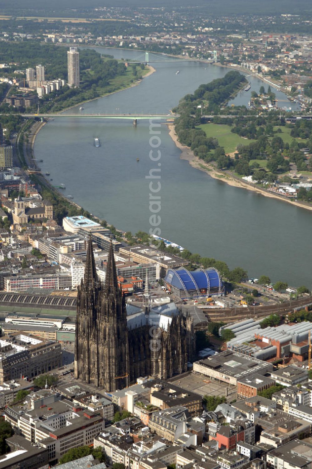 Aerial photograph KÖLN - Der Kölner Dom, offizieller Name Hohe Domkirche St. Peter und Maria, ist eine römisch-katholische Kirche in Köln und die Kathedrale des Erzbistums Köln.Der Kölner Dom ist mit 157 Metern Höhe nach dem Ulmer Münster die zweithöchste Kirche in Deutschland sowie die dritthöchste der Welt. Er steht an der nördlichen ehemaligen römischen Stadtgrenze in direkter Nachbarschaft des heutigen Hauptbahnhofs, der Altstadt, Hohenzollernbrücke und Museum Ludwig und ist von einer modernen Betonkonstruktion, der sogenannten Domplatte, umgeben. Vom rund 250 Meter entfernten Rhein und vom Hauptbahnhof aus ist die Lage der Kathedrale auf dem sogenannten Domhügel, rund 17 m über dem Rhein, noch zu erahnen. Der Kölner Dom ist die weltweit drittgrößte Kathedrale im gotischen Baustil (nach der Kathedrale von Sevilla und dem Mailänder Dom). Viele Kunsthistoriker sehen in ihm eine einmalige Harmonisierung sämtlicher Bauelemente und des Schmuckwerks im Stil der mittelalterlich-gotischen Architektur verwirklicht. Der Kölner Dom wurde 1996 in die Liste des Weltkulturerbes aufgenommen. Die riesige Fläche der Westfassade mitsamt den beiden Türmen von über 7.100 Quadratmetern ist bis heute nirgendwo übertroffen worden. Von 1880 bis 1884 war er das höchste Gebäude der Welt. Er ist zudem die populärste Sehenswürdigkeit Deutschlands: 2001 wurden fünf Millionen, 2004 sechs Millionen Besucher aus aller Welt gezählt.