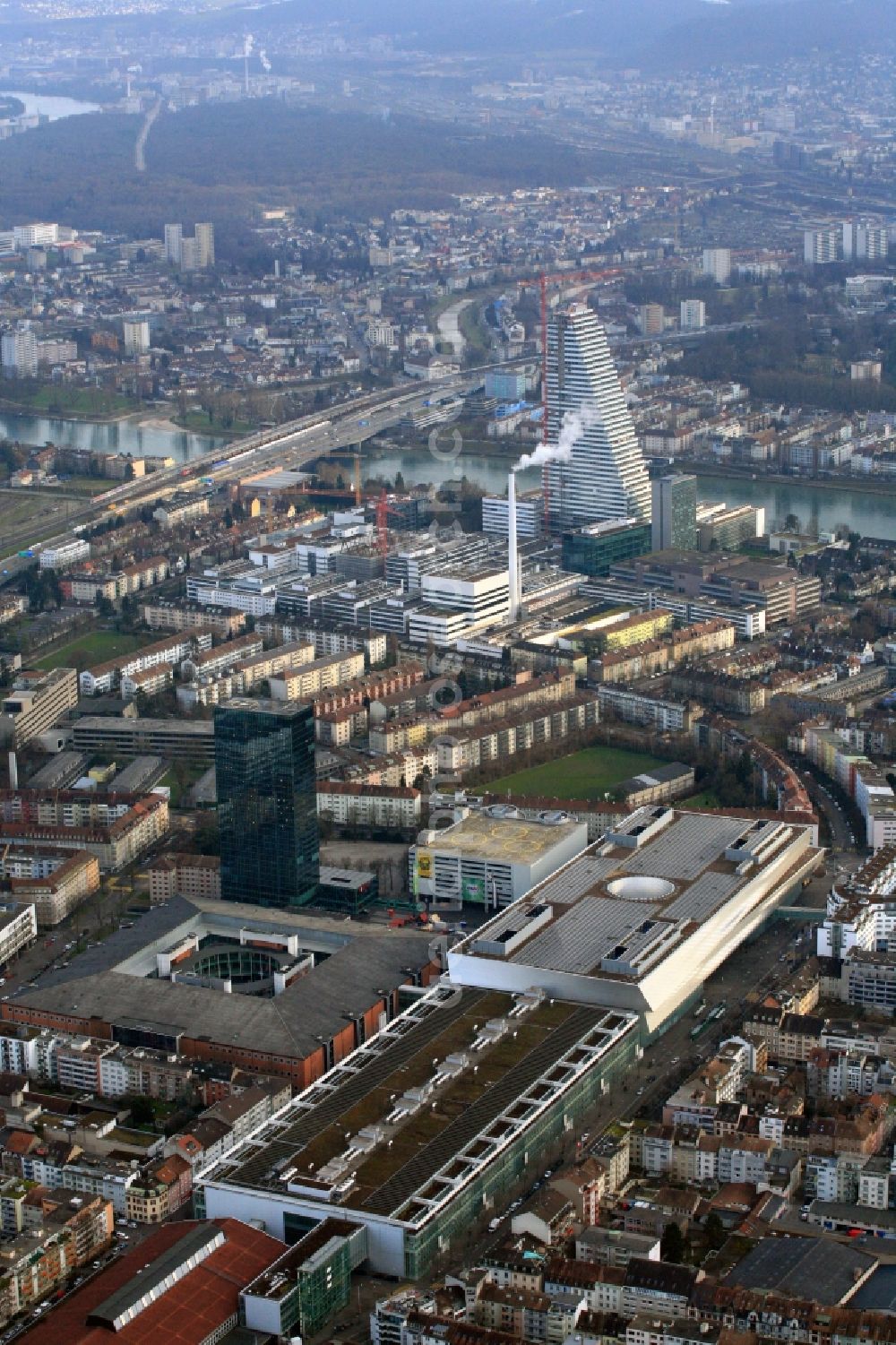 Aerial photograph Basel - In front the exhibition center Basel with the Basel Exhibition Tower, followed by the under construction Roche-tower on the premises of the pharmaceutical company Hoffmann-La Roche AG, Basel, Switzerland.Roche Tower on the premises of the pharmaceutical company Hoffmann-La Roche AG, Basel, Switzerland