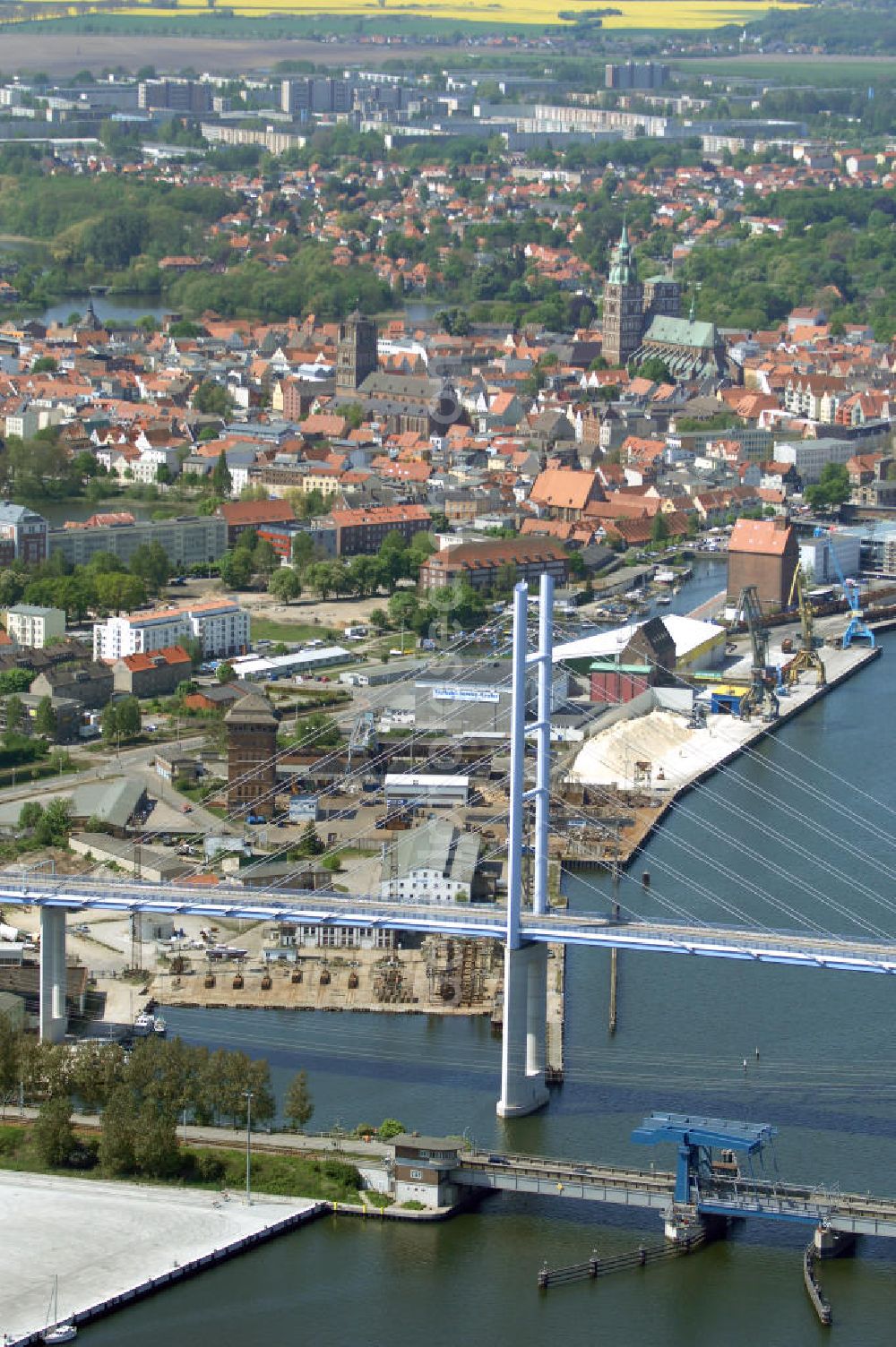 Stralsund from above - Mit dem Begriff Strelasundquerung werden die beiden Brückenverbindungen der Insel Rügen über den Strelasund zum vorpommerschen Festland bei Stralsund, die Rügenbrücke und der Rügendamm, sowie die regelmäßig betriebenen Fährverbindungen zwischen Stralsund und Altefähr sowie Stahlbrode und Glewitz bezeichnet. Der Rügendamm ist die erste feste Strelasundquerung, über den sowohl die alte Bundesstraße 96, als auch eine eingleisige Eisenbahnstrecke sowie ein kombinierter Fuß- und Radweg führen. Sie wurde 1936/1937 fertiggestellt. Rügenbrücke ist der Name der im Jahr 2007 fertiggestellten dreispurigen Hochbrücke ausschließlich für den Kraftfahrzeugverkehr zwischen der Ortschaft Altefähr auf Rügen und der Hanse- und Weltkulturerbestadt Stralsund im Zuge der als Ortsumgehung ausgebauten Bundesstraße 96. Beide Brücken werden parallel betrieben. (DEGES, MAX BÖGL)