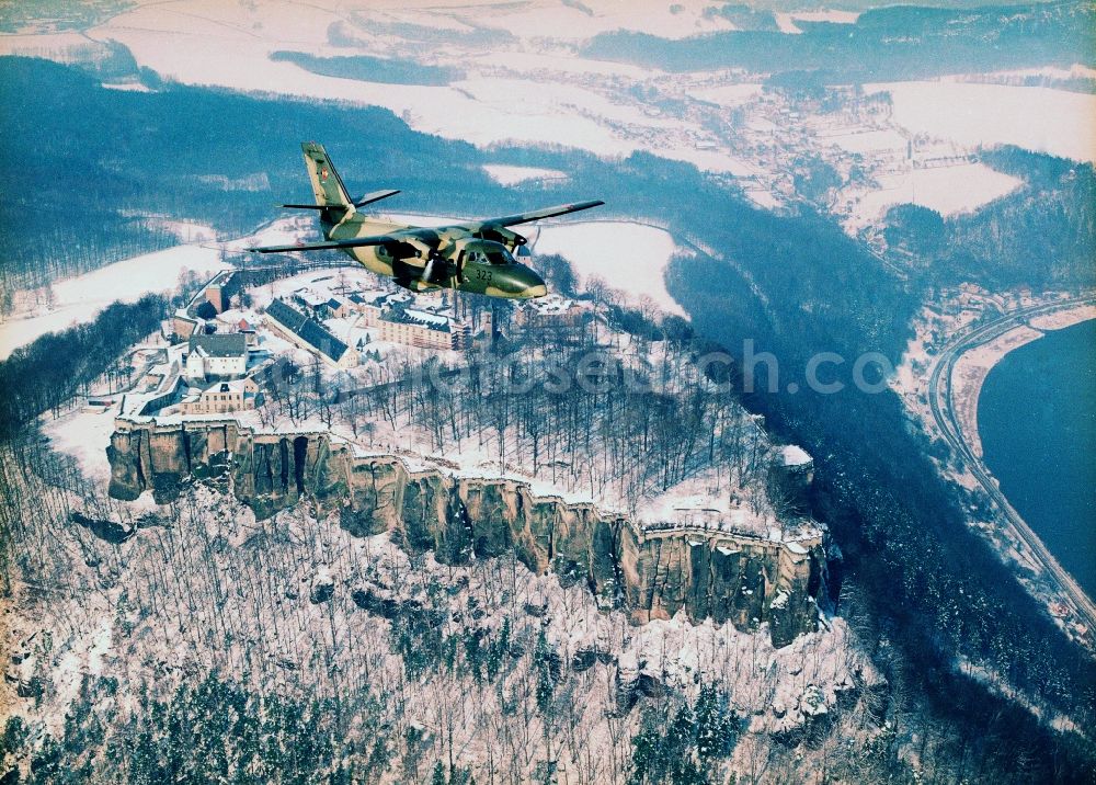 Aerial photograph Königstein - Transport and liaison aircraft L410 RRP of the NVA with the tactical marking 323 in flight over the wintry snowy Fortress Koenigstein at the river Elbe in the county district of Saxon Switzerland East Erzgebirge in the state of Saxony. The fortress is one of the largest mountain fortresses in Europe and is located amidst the Elbe sand stone mountains on the flat top mountain of the same name. In front of it, the river runs through a valley